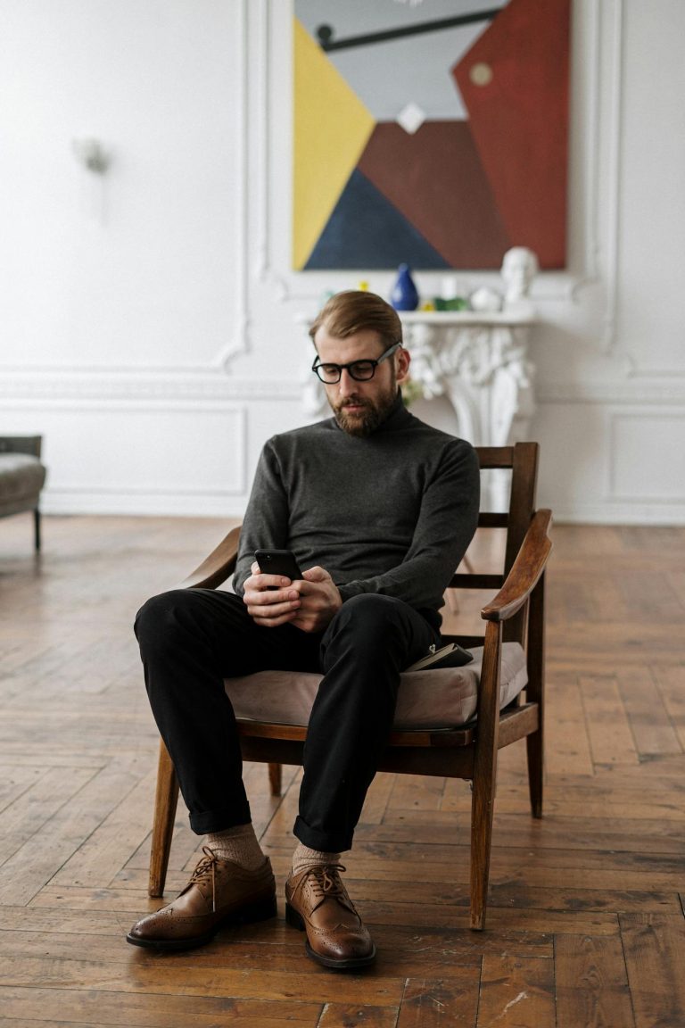 Stylish man in glasses seated on wooden armchair, checking smartphone in contemporary interior.